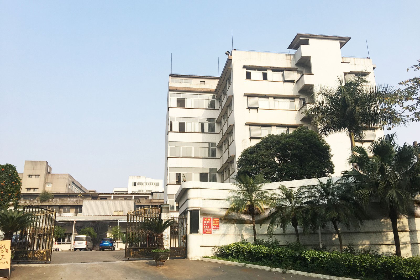 Multi-story factory building exterior with palm trees and entrance gate