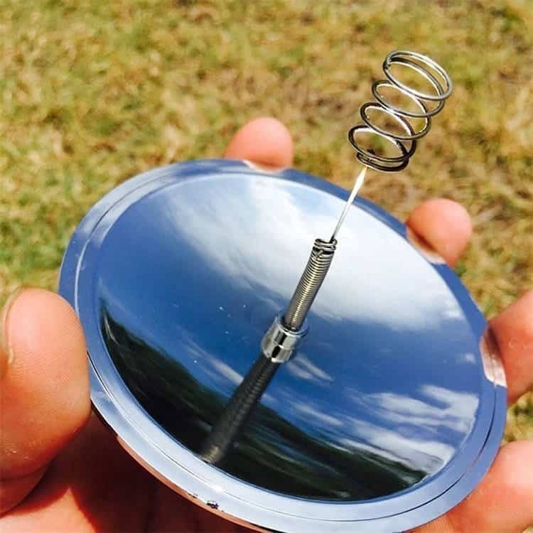 Hand holding a 3.8-inch silver solar spark lighter from the ferro rod bulk FRC29 line, concentrating sunlight to ignite tinder on a sunny day.